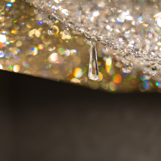 Luxury Rectangular Crystal Chandelier Close-up of a sparkling Luxury Rectangular Crystal Chandelier with numerous hanging crystals. One long, teardrop-shaped crystal is prominently in focus, reflecting light and creating a spectrum of colors against a blurred background.