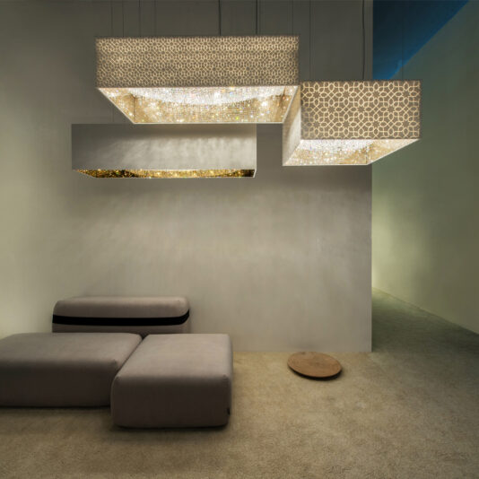 A minimalist interior features a luxury rectangular crystal chandelier with intricate, textured surfaces hanging from the ceiling. Below are two light gray, low-profile sofas on a beige carpeted floor, creating a cozy, modern lounge space.
