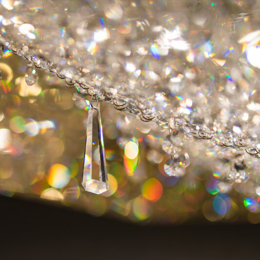 Luxury Square Crystal Chandelier Close-up of the Luxury Square Crystal Chandelier showcasing a dangling crystal surrounded by shimmering and blurred multicolored lights. The image captures the sparkle and reflective beauty of the chandelier's crystals against a warm, bokeh-filled background.