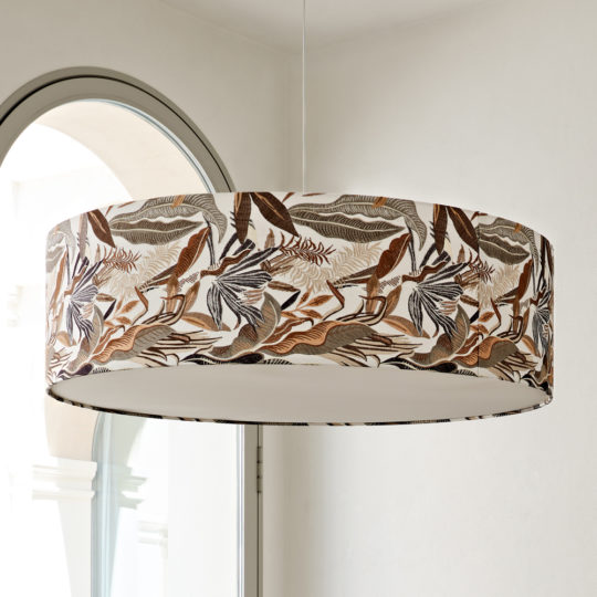 A Luxury Suspension Light With Embroidered Shade, adorned with a tropical leaf pattern in shades of brown and beige, hangs near an arched window, allowing natural light to fill the room. The backdrop of the white walls enhances its elegance.