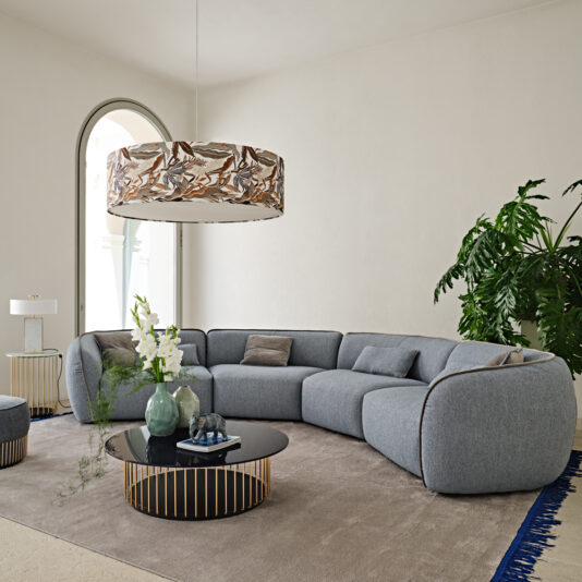 Luxury Suspension Light With Embroidered Shade A modern living room featuring a large, curved gray sofa adorned with throw pillows, a round black coffee table with a gold base, and the Luxury Suspension Light With Embroidered Shade overhead. The room has a light-colored rug, a large plant, and an arched doorway in the background.