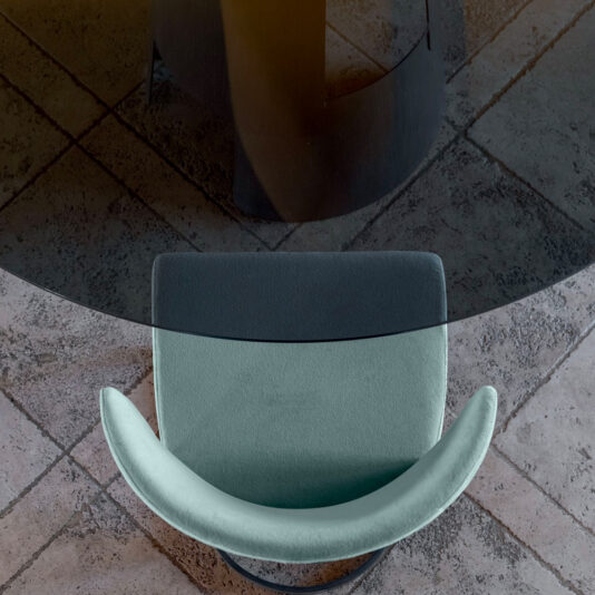 A modern chair with a mint green cushion is positioned next to a round glass table on a tiled floor. The perspective is from above, showing the curvature of the chair's backrest and the reflection of the table's base in the glass surface.