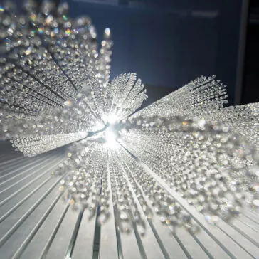 Luxury Vertical Crystal LED Chandelier A close-up of the Luxury Vertical Crystal LED Chandelier reveals a shimmering, abstract glass sculpture with crystal-like rods radiating outward. Sunlight creates bright reflections and a prismatic effect against the striped backdrop.
