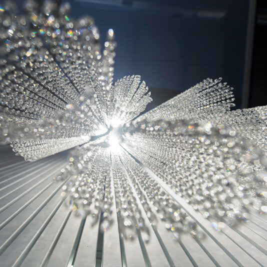 A close-up of the Luxury Vertical Crystal LED Chandelier reveals a shimmering, abstract glass sculpture with crystal-like rods radiating outward. Sunlight creates bright reflections and a prismatic effect against the striped backdrop.