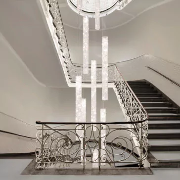 Luxury Vertical Crystal LED Chandelier A grand staircase with ornate white railings and black steps features a modern Luxury Vertical Crystal LED Chandelier cascading from a high ceiling, softly illuminating the elegant architectural details. The walls and ceiling are painted white, offering a clean, sophisticated look.