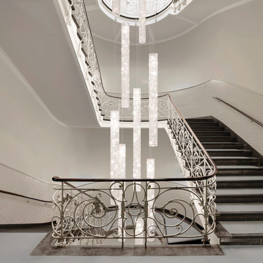 A grand staircase with ornate white railings and black steps features a modern Luxury Vertical Crystal LED Chandelier cascading from a high ceiling, softly illuminating the elegant architectural details. The walls and ceiling are painted white, offering a clean, sophisticated look.