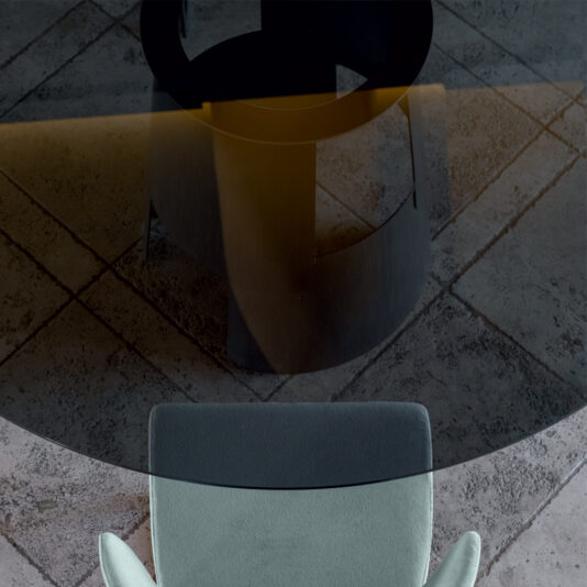 Modern Round Glass Dining Table A top-down view of the Modern Round Glass Dining Table featuring a sleek black base and a light gray upholstered chair positioned on a tiled floor. The glass tabletop exhibits a subtle reflective sheen, creating an interplay of light and shadow.