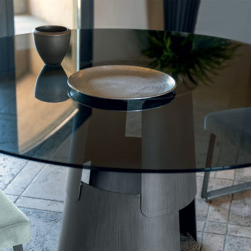 Modern Round Glass Dining Table A Modern Round Glass Dining Table featuring a decorative ceramic cup and a textured plate on top. The table is set on a tiled floor, with a chair visible in the foreground and a plant partially visible in the background.