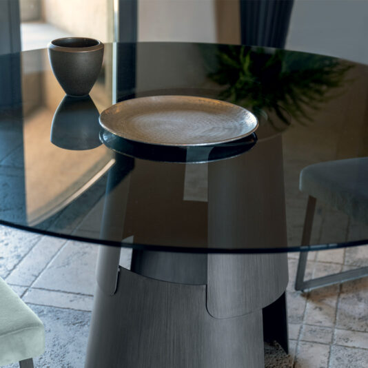 A Modern Round Glass Dining Table featuring a decorative ceramic cup and a textured plate on top. The table is set on a tiled floor, with a chair visible in the foreground and a plant partially visible in the background.
