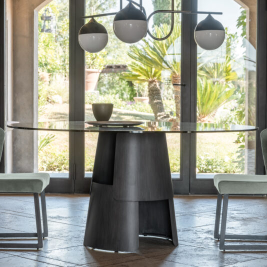 A modern dining area features the sleek Modern Round Glass Dining Table, supported by a sculptural dark base. This contemporary centerpiece is flanked by two chairs with light-colored upholstery. Above, a stylish pendant light with three bulbs hangs. Large glass doors in the background showcase a lush garden.