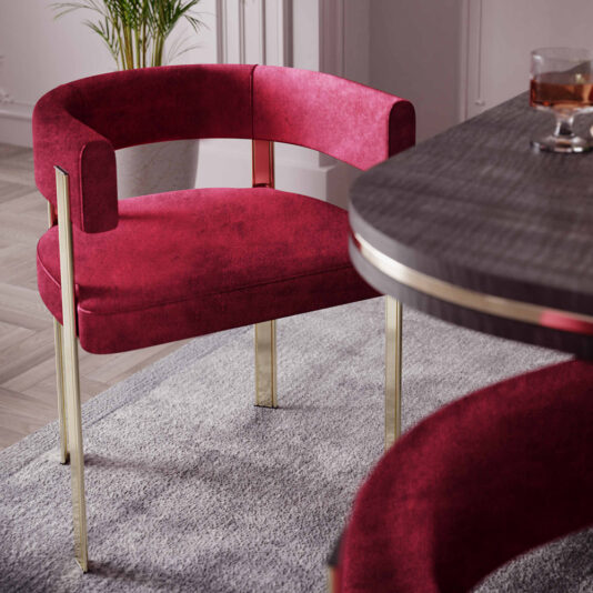 The Modern Curved Dining Chair, featuring plush red upholstery and gold metal legs, stands prominently on a light gray rug. The chair, with its round back design, is placed near a dark wood dining table accented with gold trim. The room boasts light walls and parquet flooring, while a plant is visible in the background.