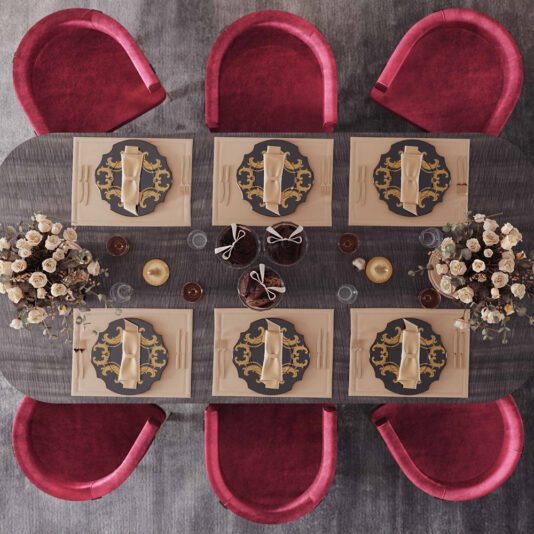 Modern Curved Dining Chair A top view of a rectangular dining table set for six with elegant tableware, including decorative plates, beige napkins, and cutlery. Floral centerpieces with white and blush roses adorn the table. The table is surrounded by six Modern Curved Dining Chairs upholstered in red velvet.