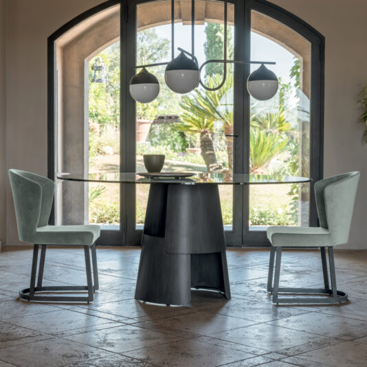 A modern dining area showcases a Modern Italian Dining Set, which includes a round glass table with a striking black, sculptural base, flanked by two light green upholstered chairs. Overhead, a trio of contemporary pendant lights illuminates the space. Large arched windows in the background offer a picturesque view of the lush garden outside.