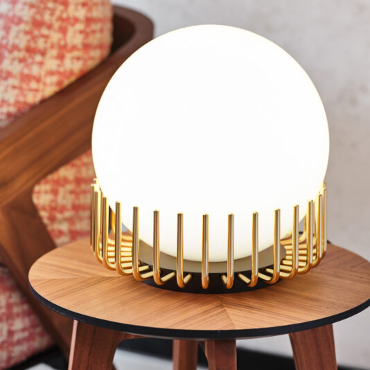 A **Small Contemporary Sphere Table Lamp**, radiating a warm white glow, is elegantly housed in a golden slatted metal frame, resting on a wooden table. Accompanying this setup is a red and white patterned, cushioned chair against a textured wall background, creating a modern yet cozy ambiance.