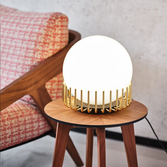 Small Contemporary Sphere Table Lamp The Small Contemporary Sphere Table Lamp features a modern design with a round, frosted glass shade encased in a stylish gold metal frame. It sits gracefully on a wooden table with a tripod base, while in the background, a red and white textured chair is partially visible against a light-colored wall.