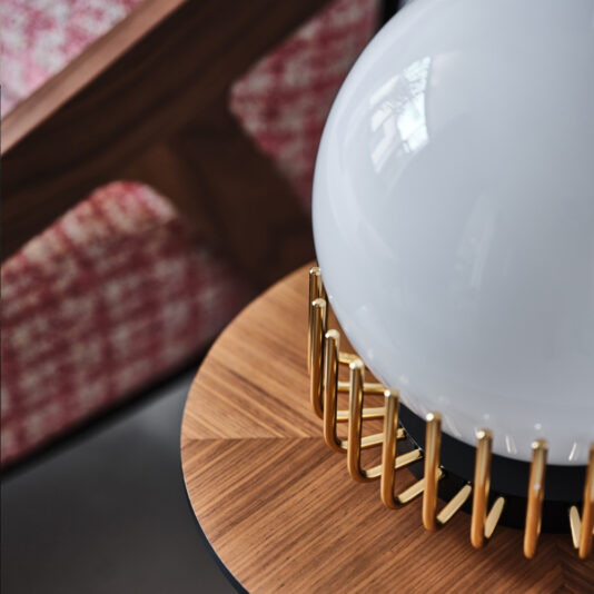 Small Contemporary Sphere Table Lamp A Small Contemporary Sphere Table Lamp with a gold metallic base sits on a wooden table featuring a geometric pattern. In the background, a red and white patterned cushion rests on a wooden chair.