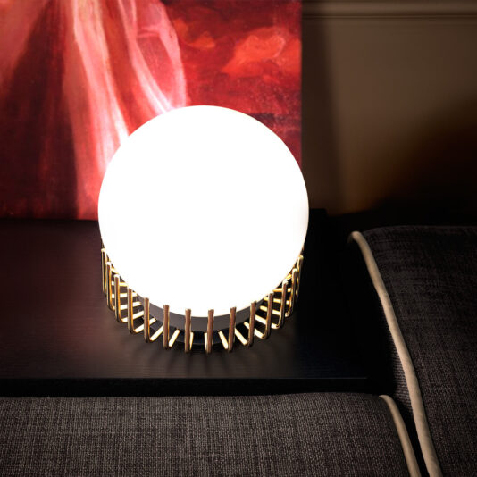 Small Contemporary Sphere Table Lamp The Small Contemporary Sphere Table Lamp, featuring a gold, cage-like base, is perched on a tabletop. Emitting a bright light, it illuminates part of a dark-colored sofa accented with light piping in the background. Set before a large, reddish abstract painting, this lamp adds a touch of modern flair to the space.