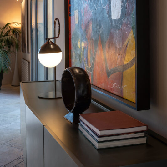 A Smoked Black Ash Sideboard hosts a sleek lamp, a stack of hardcover books, and a unique black sculpture. Behind it hangs an abstract painting. Light from a nearby window illuminates the scene, highlighting the contemporary decor.