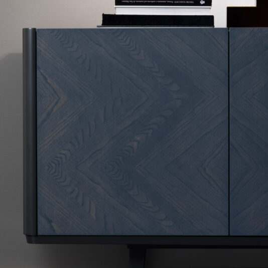 Close-up of the Smoked Black Ash Sideboard showcasing a striking chevron pattern on the door. The intricate wood grain is accentuated by the angular design, giving it a sophisticated and modern appearance. A small portion of objects on top of the sideboard is barely visible.