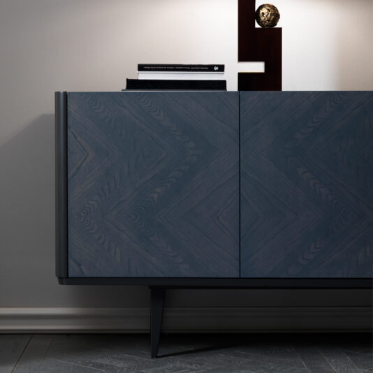 A close-up view of the Smoked Black Ash Sideboard featuring a chevron pattern. The top of the sideboard is adorned with several stacked books and a sculpture. The background wall is painted light grey, and part of the scene is illuminated by a light.