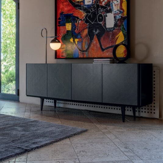 A modern room featuring the Smoked Black Ash Sideboard with angled legs, decorated with a geometric lamp and a circular frame. Above the sideboard hangs a colorful abstract painting. The room has tiled flooring, a grey rug, and a large window partially showing greenery outside.