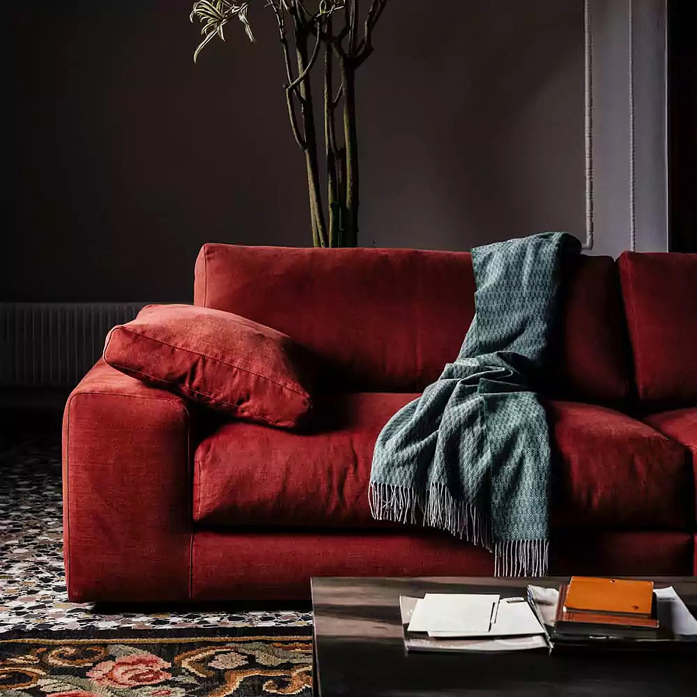 A cozy living room scene features a red sofa adorned with a folded green throw blanket and matching cushion. A dark wooden coffee table in the foreground holds a stack of books, a magazine, and a closed journal—perfect for chasing away those Blue Monday blues. A tall plant stands behind the sofa.