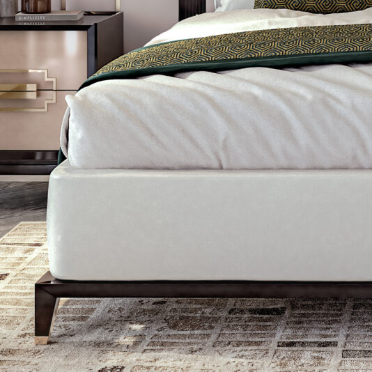 Art Deco Style Bed With Tall Headboard Close-up of a modern bedroom corner featuring the Art Deco Style Bed With Tall Headboard, complete with an upholstered white base and covered in white bedding with a green and gold-patterned blanket. Next to it, a nightstand with a drawer and metallic handles holds a small lamp on top. A geometric-patterned rug adorns the floor.