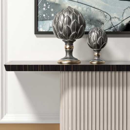 A minimalist interior features a black floating shelf adorned with two metallic artichoke sculptures of varying sizes. Just below, the Art Deco Style Veneer Console Table lends an air of sophistication to the space. Behind the shelf, a framed abstract painting in cool tones is mounted on a white wall with vertical paneling beneath the shelf.