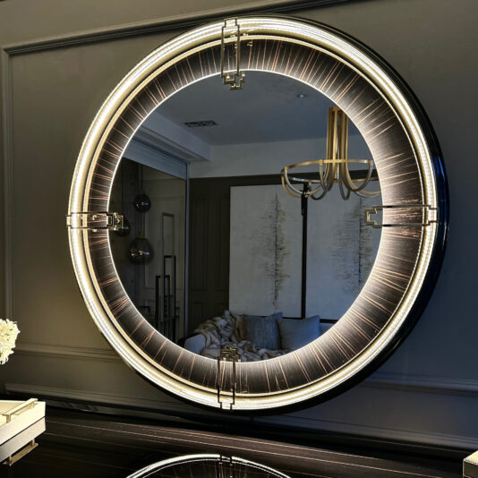 The Backlit Art Deco Style Veneer Round Mirror, with its large, round design and thick backlit frame, is mounted on a wall. It reflects part of a modern, elegantly decorated room featuring a grey wall, abstract paintings, a chandelier, and a glimpse of a sofa.