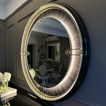Backlit Art Deco Style Veneer Round Mirror The Backlit Art Deco Style Veneer Round Mirror with its sleek black frame hangs on the wall. This round mirror, featuring embedded LED lights, creates a glowing halo effect. In its reflection, you can see a dining room adorned with white chairs, a table, and a floral centerpiece.