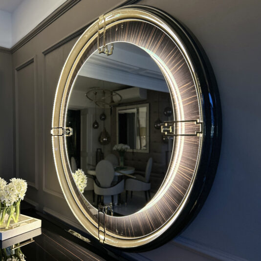 The Backlit Art Deco Style Veneer Round Mirror with its sleek black frame hangs on the wall. This round mirror, featuring embedded LED lights, creates a glowing halo effect. In its reflection, you can see a dining room adorned with white chairs, a table, and a floral centerpiece.