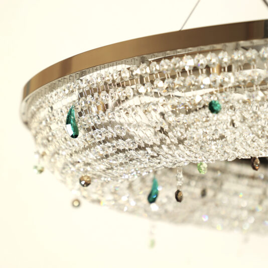 A close-up view of the Contemporary Crystal Chandelier With Coloured Drops showcases its metallic frame adorned with numerous crystal beads. The chandelier is accentuated by a few green and dark glass droplets, which add a touch of color to the sparkling design. The plain white background highlights the chandelier's intricate details.