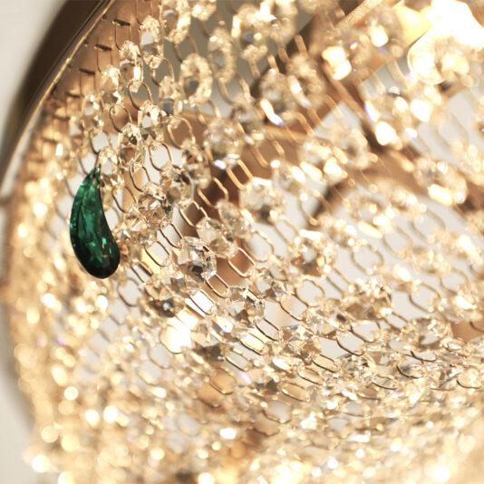 Close-up image of a Contemporary Crystal Chandelier With Coloured Drops, featuring intricate gold metalwork and numerous sparkling crystals. Among the clear drops, a single large green crystal stands out, creating a striking contrast.