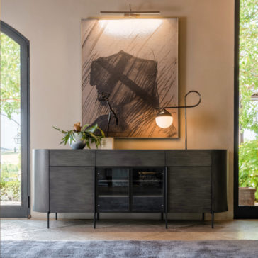 Contemporary Sideboard With Glass Doors A modern living room showcases a dark wood console table with curved edges, beneath which a large abstract painting is illuminated by a picture light. The *Contemporary Sideboard with Glass Doors* holds decorative items like a potted plant and a sleek lamp, while large windows reveal beautiful greenery outside.