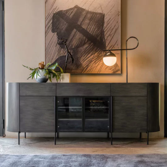 A Contemporary Sideboard With Glass Doors with a dark wood finish is placed against a beige wall. Atop the sideboard is a sculptural lamp featuring a curved stem and a round white shade, along with a gray vase containing green plants, and an oversized framed abstract art piece. A textured rug lies in front.