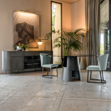 Contemporary Sideboard With Glass Doors A modern dining room featuring a round glass table with a black base, surrounded by two sage green upholstered chairs with black legs. A large contemporary artwork hangs above a Contemporary Sideboard With Glass Doors, while plants and a table lamp add to the decor. Floor-to-ceiling windows with beige curtains let in natural light.