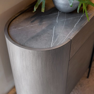 Contemporary Sideboard With Glass Doors Introducing the Contemporary Sideboard With Glass Doors: a modern piece featuring softly rounded edges and a sophisticated dark grey marble top with striking white veining. It’s beautifully complemented by a metal pot with lush green foliage. The sides boast a stylish brushed metal finish, while the elegant glass doors provide a glimpse at your stored treasures inside.