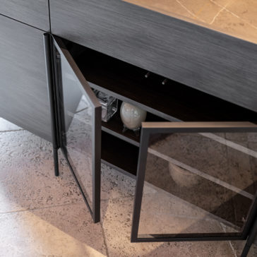 Contemporary Sideboard With Glass Doors A close-up of a Contemporary Sideboard With Glass Doors in a modern kitchen with a grey wood finish. The open cabinet door reveals shelves inside, containing a large light bulb and various stored items. The flooring features textured stone tiles.