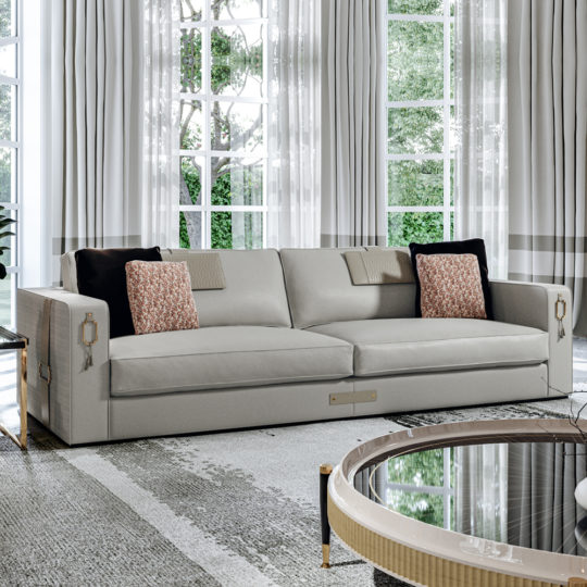 A modern living room features the High End Art Deco Style Sofa adorned with black and patterned throw pillows. The elegant sofa faces a round coffee table with a glass top and light wood trim. Floor-to-ceiling windows with sheer curtains showcase views of a green, leafy garden outside.