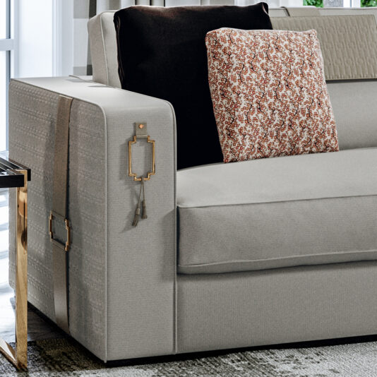 High End Art Deco Style Sofa A sophisticated beige High End Art Deco Style Sofa, adorned with a black cushion and a patterned throw pillow, boasts an armrest embellished with decorative stitching and a unique hanging key-shaped accessory. Positioned adjacent to the sofa is a sleek glass and metal side table, as light streams in through a nearby window.