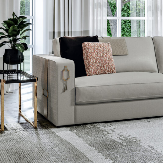 High End Art Deco Style Sofa A modern living room showcases a High End Art Deco Style Sofa in light gray, decorated with two pillows—one black and one featuring a floral pattern. An elegant side table with a gold frame and a sleek black top supports a potted plant. Large windows in the background offer views of lush greenery outside.