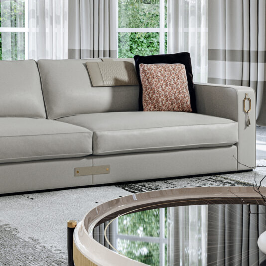 High End Art Deco Style Sofa A stylish, contemporary living room features a **High End Art Deco Style Sofa** adorned with a beige throw and a decorative pillow with a floral pattern. In front of the sofa is a round, glass-top coffee table. The room is illuminated by natural light from large windows behind the sofa.