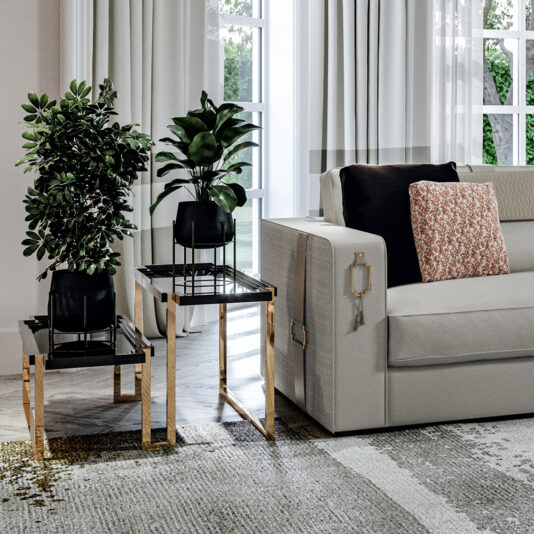 A modern living room features a light gray sofa decorated with black and patterned throw pillows. Two potted plants are placed on High End Double Tiered End Tables, which boast gold accents and black tops. Floor-to-ceiling windows draped with sheer white curtains let natural light flood the room.