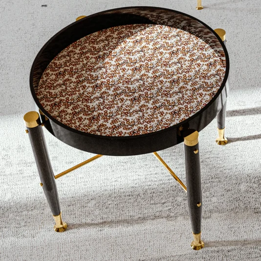 The High End Round Side Table features a patterned top with a brown and white design, four dark legs enhanced with golden accents, all set against a light carpeted floor.