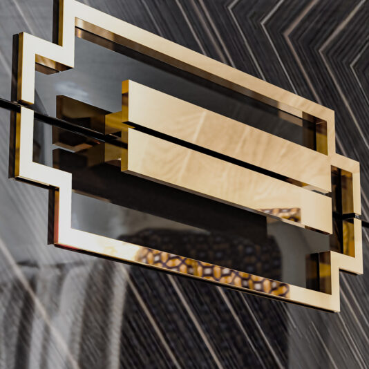 A geometric, modern light fixture with a reflective golden rectangular frame features a central horizontal bar flanked by symmetrical cut-outs on a dark, patterned wall background. Paired perfectly with the High End Veneer Bedside Cabinet, the design creates an elegant and contemporary aesthetic.