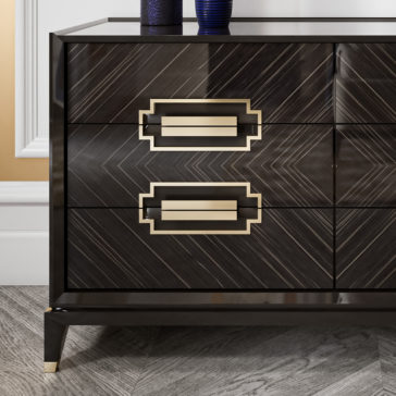 High End Veneer Chest Of Drawers A close-up of the High End Veneer Chest Of Drawers showcases its modern, dark-toned finish and sleek geometric patterns. The drawer handles are rectangular with elegant gold accents, while the chest stands on pointed legs featuring gold tips. It is positioned against a neutral white-paneled wall and rests on a parquet floor.