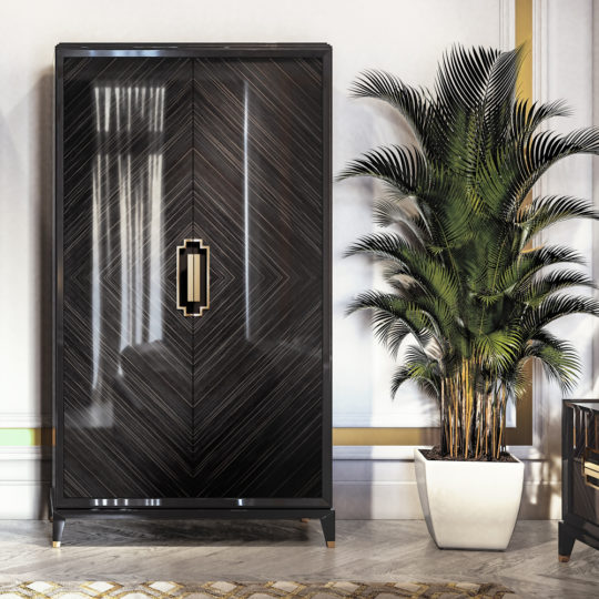 The High End Veneer Wardrobe Cabinet, featuring a sleek, modern black design and an intricate chevron pattern, stands on a marble floor. Luxurious gold handles enhance its elegance, while to its right, a tall potted plant with lush green fronds brings life to the sophisticated setting.
