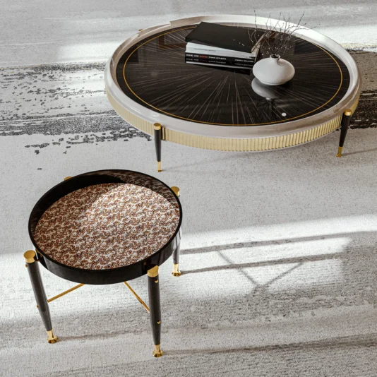 High End Veneered Low Round Coffee Table The High-End Veneered Low Round Coffee Table, with its dark wooden top, displays books and a small twig vase. A nearby smaller round table with a patterned top and gold accents stands on a light textured floor, both casting subtle shadows.