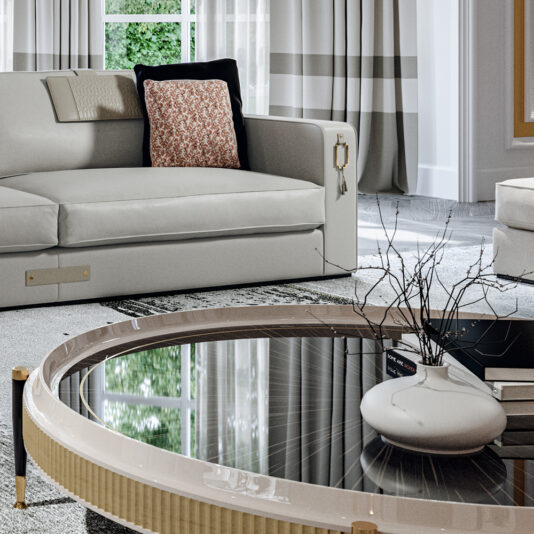 High End Veneered Low Round Coffee Table A modern living room features a white sofa adorned with cushions, one with a floral pattern. In front of the sofa is the sleek *High End Veneered Low Round Coffee Table* holding a white vase with bare branches. The room is brightly lit with natural light from a large window.