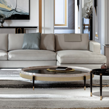 Large Chaise Art Deco Style Sofa A modern living room showcasing the Large Chaise Art Deco Style Sofa in light gray, adorned with a blue-green throw pillow. In front of the sofa sits a round coffee table featuring a glass top and gold accents. The room maintains a neutral color palette, complemented by art and décor on the walls.